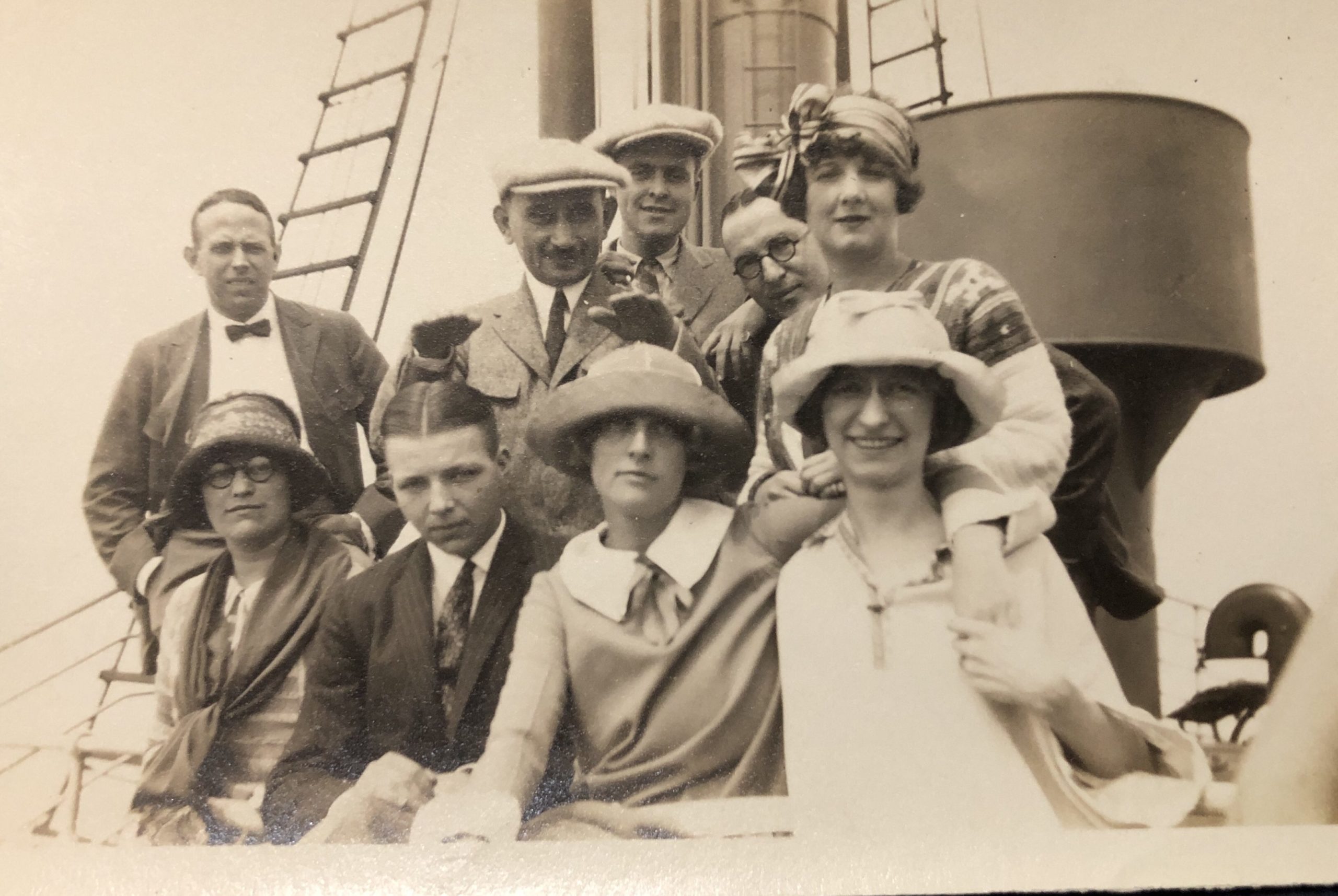 Nine people (they appear to be four women, five men) pose for a photo. The appear to be in front of a vent or steam chimeny on an ocean liner. The photo was taken in 1924,